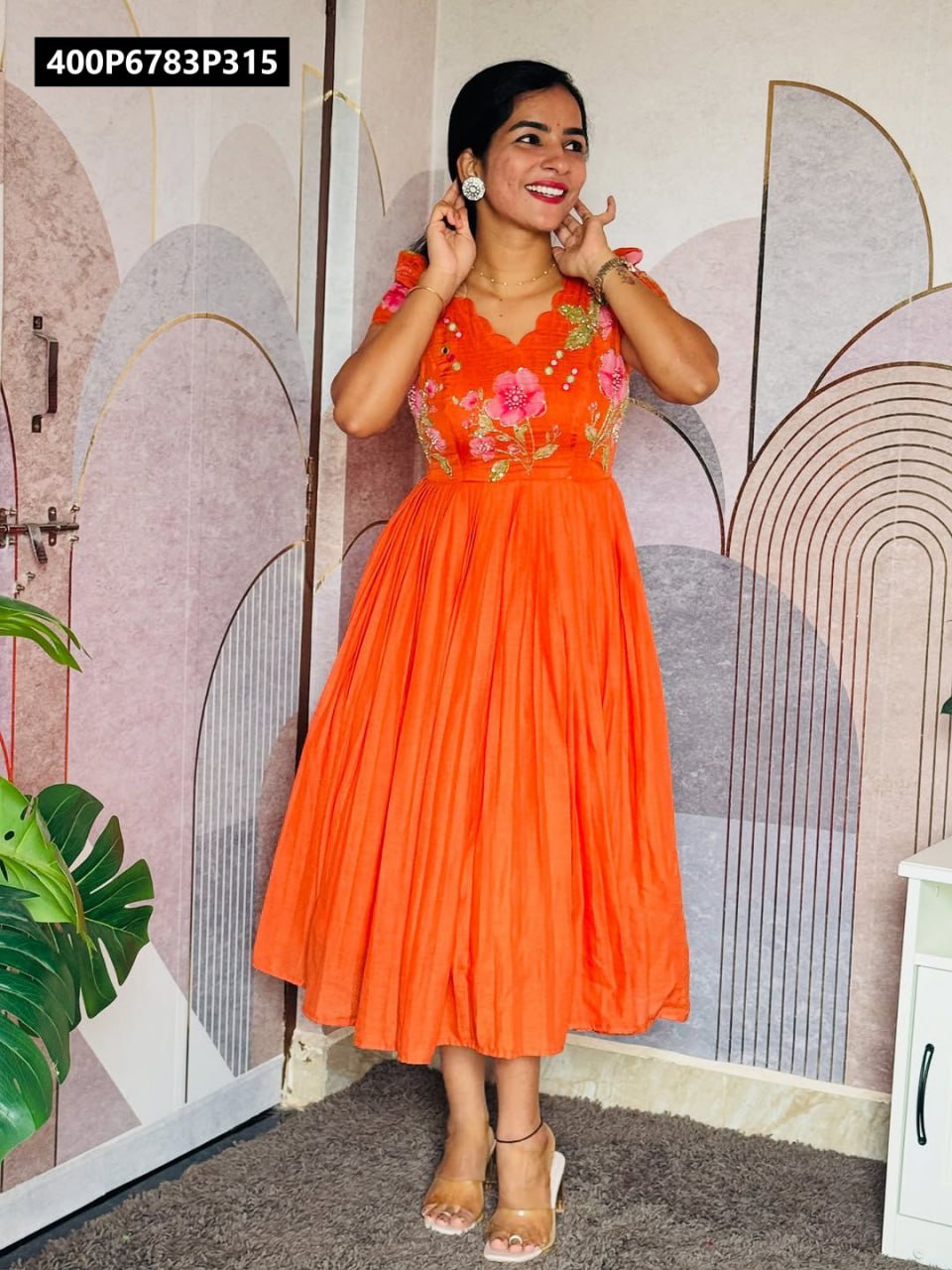 Full length shot of a smiling model in a vibrant orange TRENDBUY mul mul cotton gown with floral Maggam work on the bodice, against an artistic patterned wall.