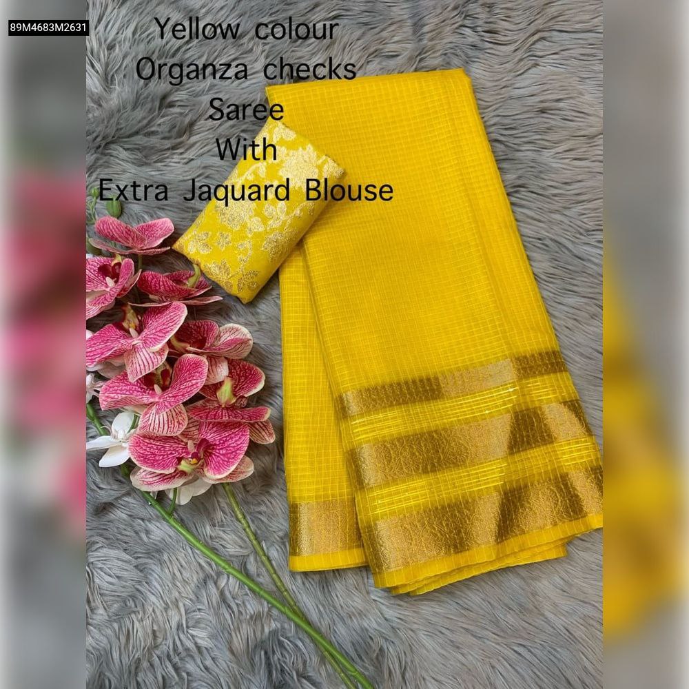 Yellow organza checks saree with golden jacquard border and matching brocade blouse piece, laid out on a grey faux fur background with decorative pink orchids.