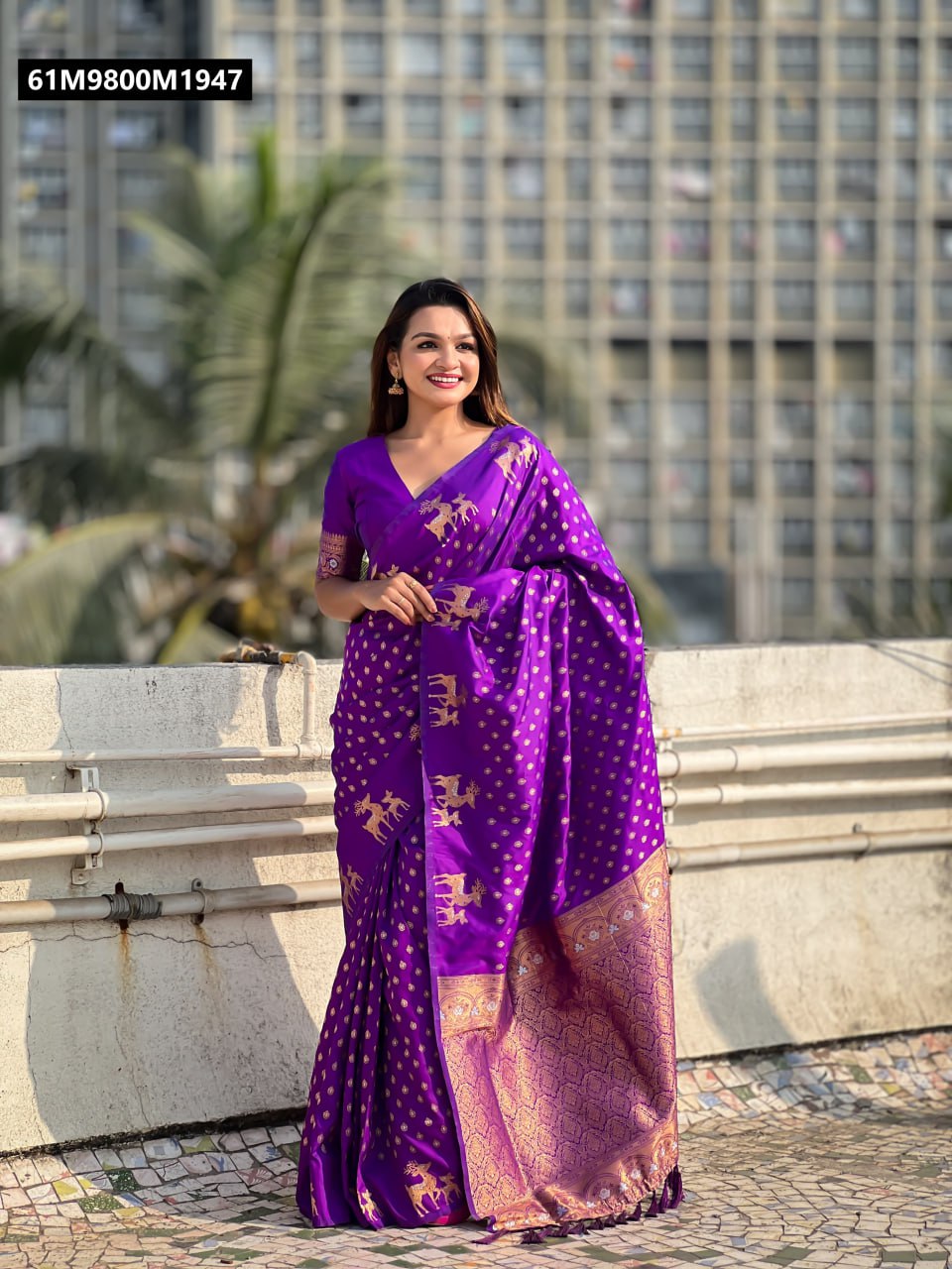 TRENDBUY woman model in a vibrant purple soft silk saree with golden zari deer motifs, full view smiling outdoors against a city backdrop.