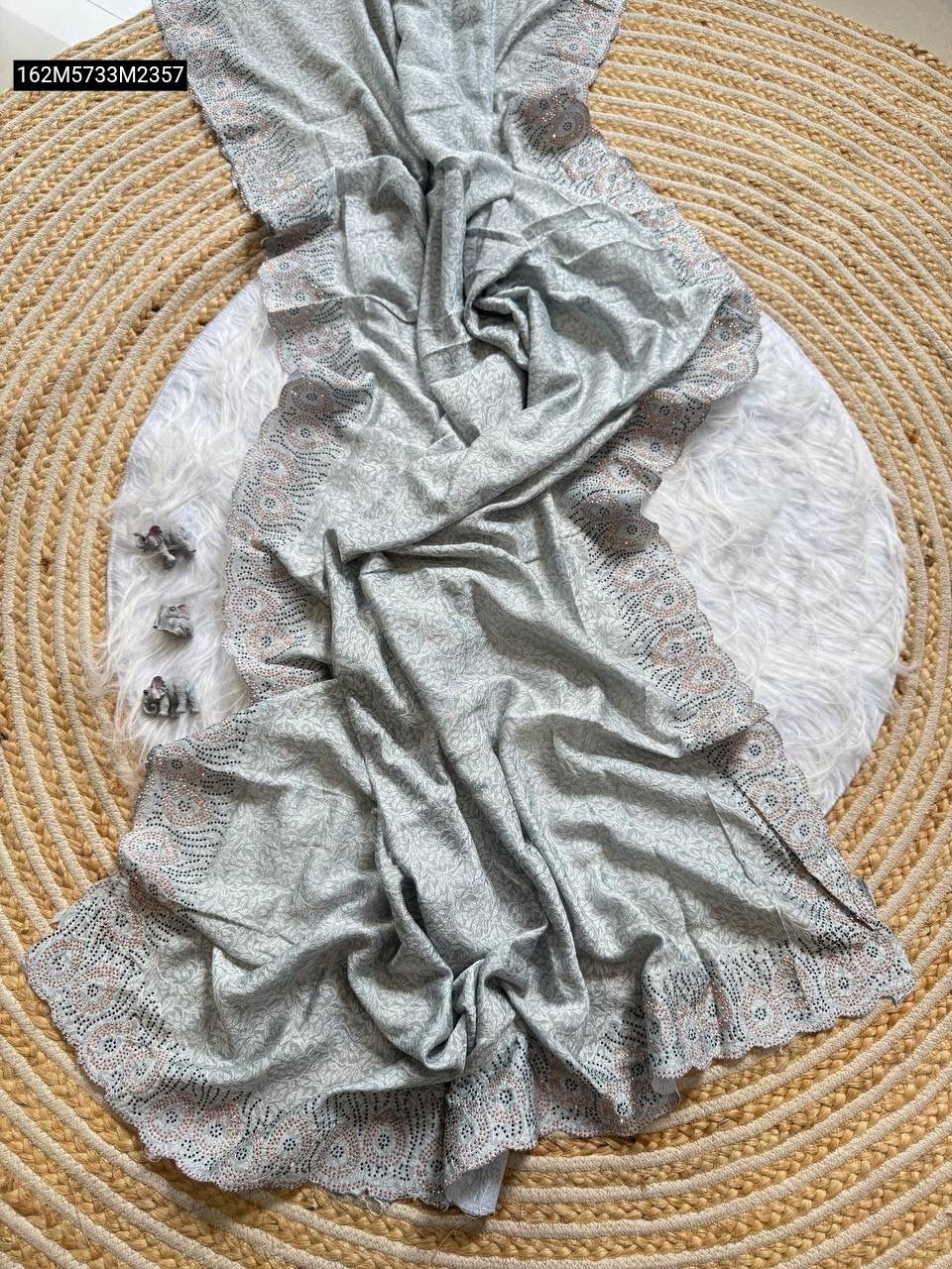 Draped Soft Grey Vichitra silk saree with subtle floral weave and ornate scalloped lace border, displayed on a rustic jute mat and fluffy white rug.