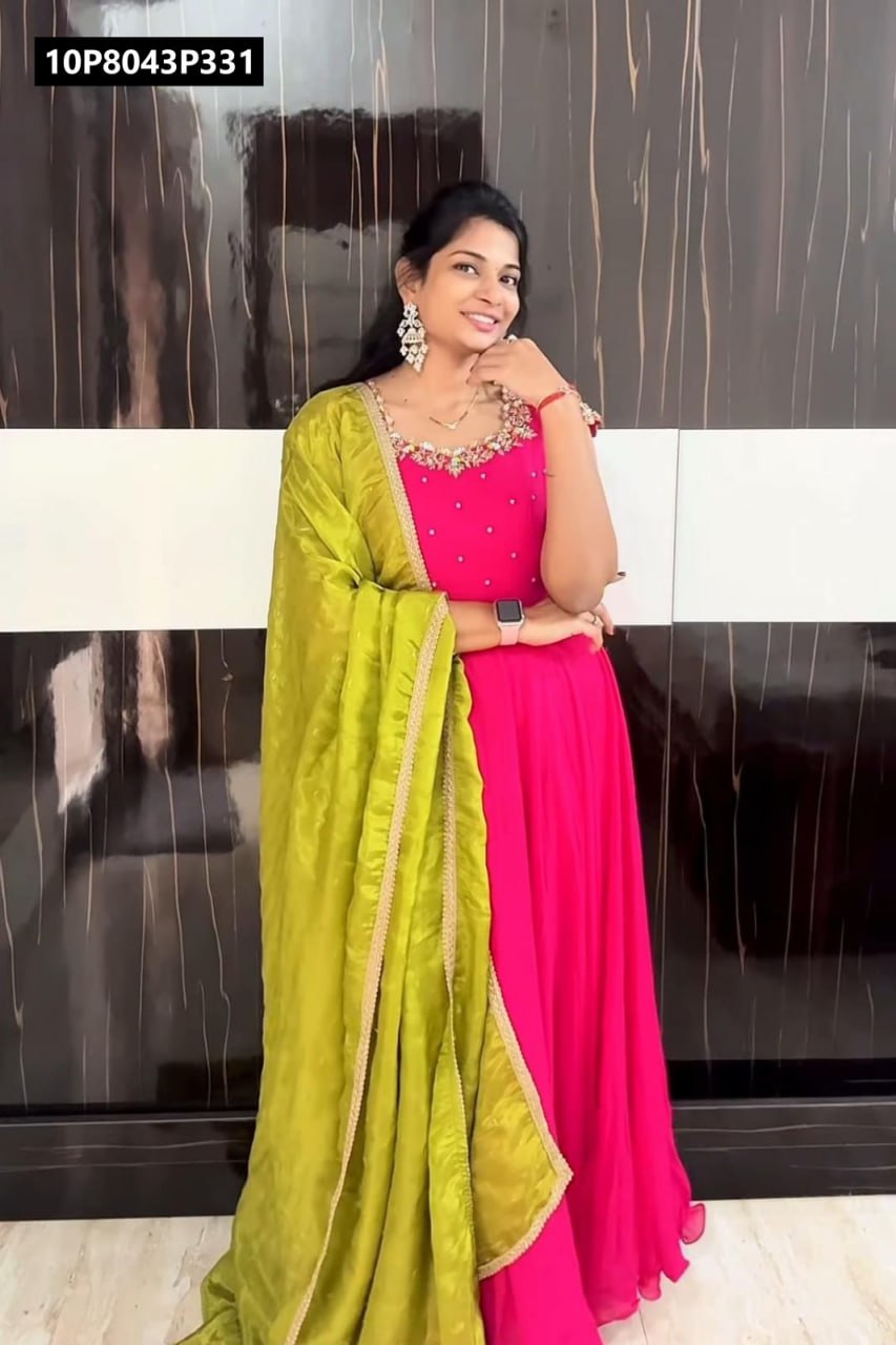 Full-length shot of a smiling Indian woman posing elegantly in a hot pink Vichitra silk Anarkali gown with ornate Maggam work, complemented by a flowing lime green Chinnon silk dupatta, against a stylish dark wood background.
