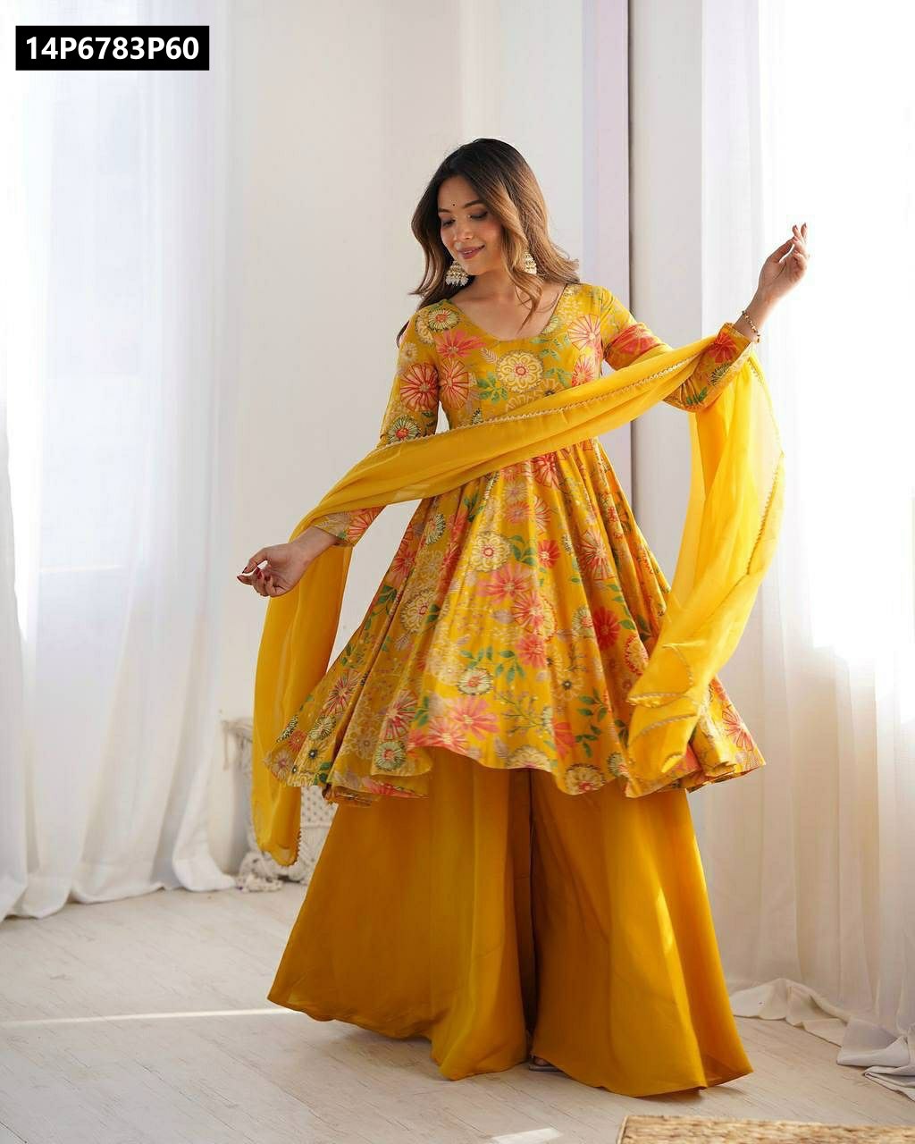 A smiling model in a full-length shot, wearing a bright yellow faux georgette floral print anarkali kurti, matching palazzo pants, and a sheer dupatta.