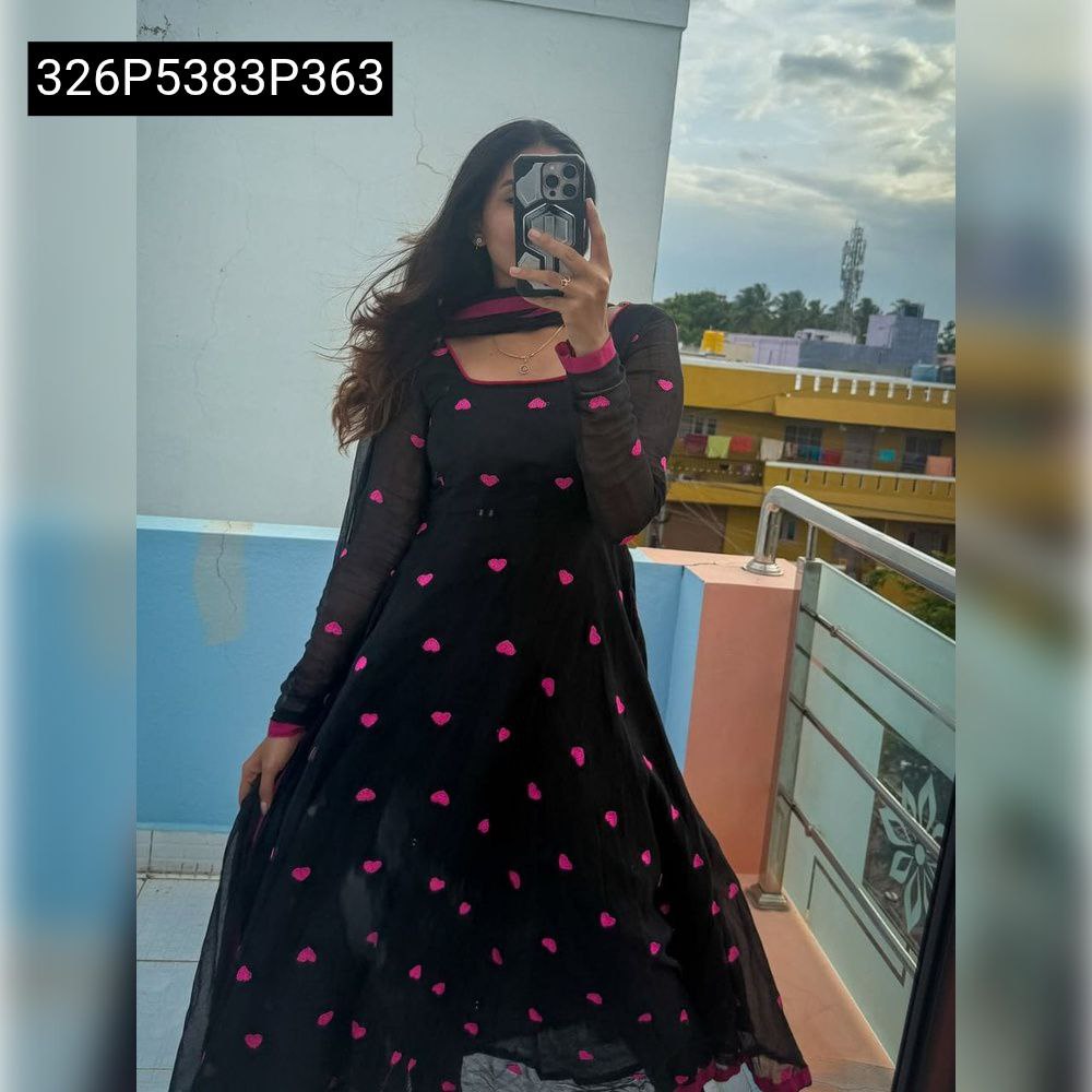 Full body shot of a model elegantly posing in a black georgette Anarkali gown with pink heart embroidery, outdoors on a balcony.