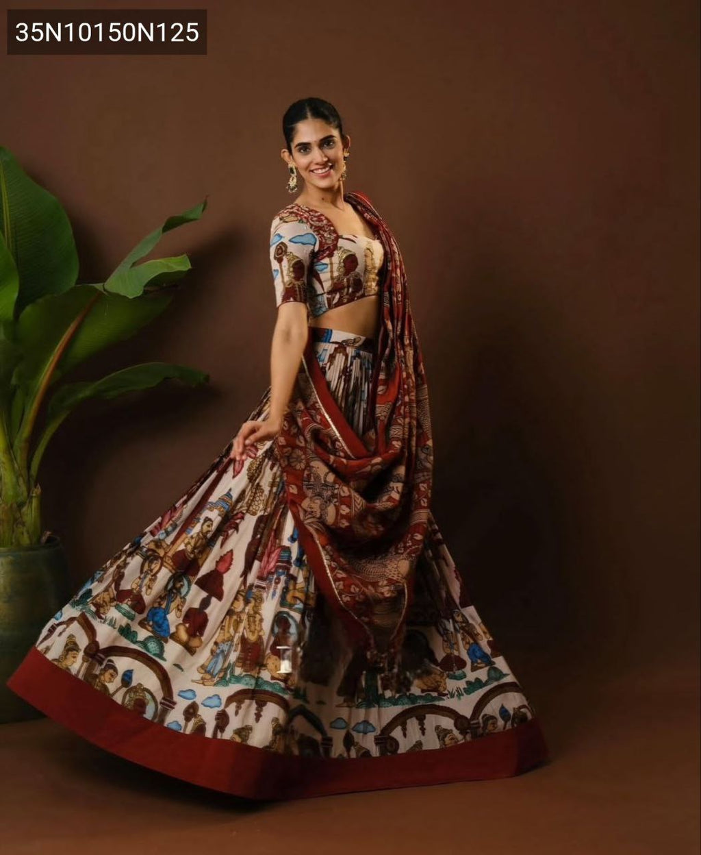 Full body shot of a smiling woman in a TRENDBUY Off-White and Maroon Kalamkari print cotton lehenga choli set, posing dynamically with a plant in the background, showcasing the full flair.