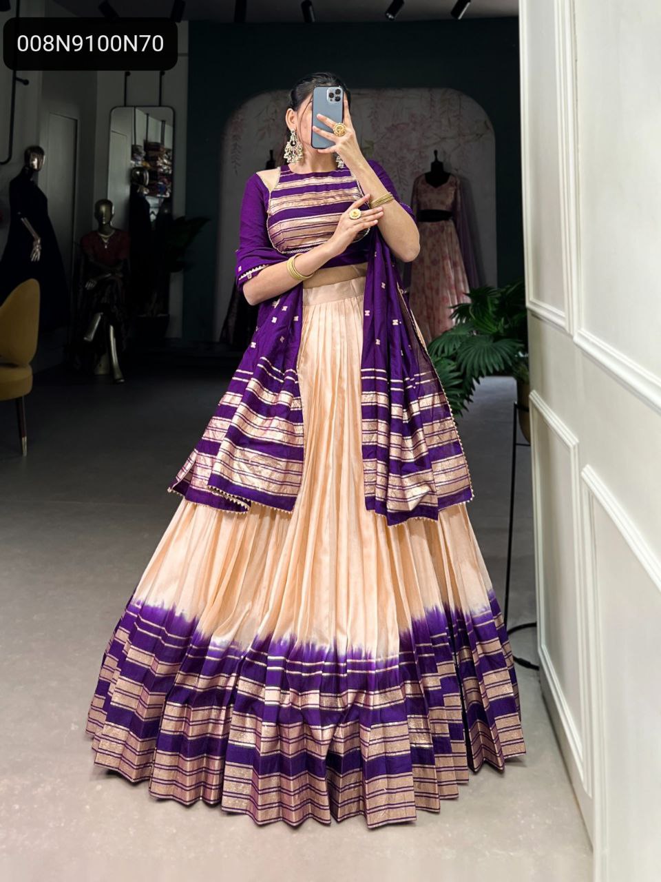 Full shot of a model wearing a peach-colored lehenga with a purple tie-dye and gold zari striped border, paired with a matching purple blouse and dupatta, posing in a mirror.