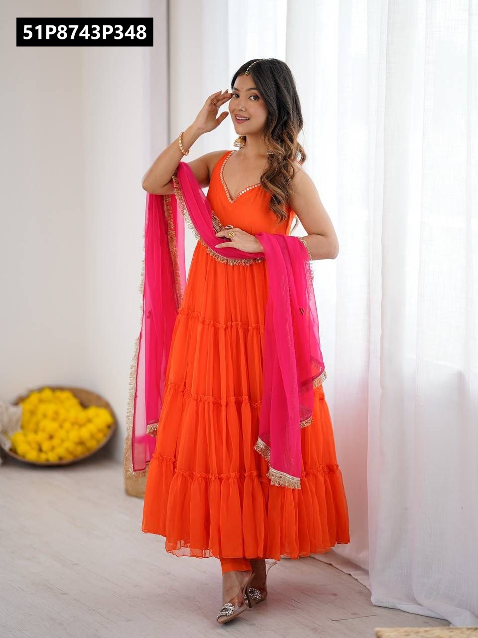 Model smiling in a sleeveless orange tiered georgette gown and hot pink dupatta with golden lace, hands near face.