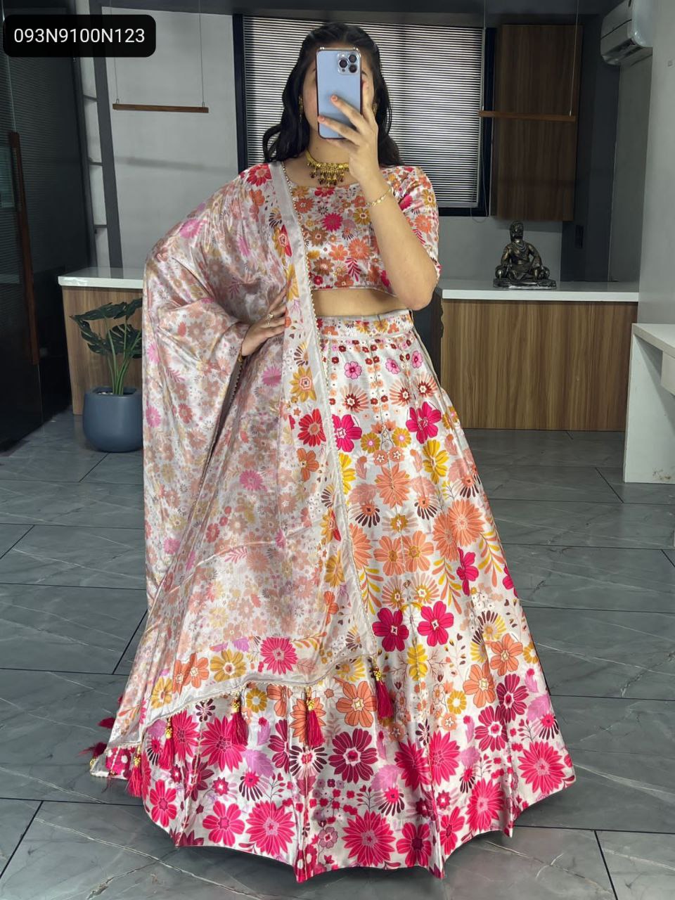 Full body shot of model wearing a white/cream TRENDBUY art silk printed lehenga choli with vibrant pink and orange floral pattern, gota patti work, and matching chinon dupatta with tassels, posed in a modern studio.