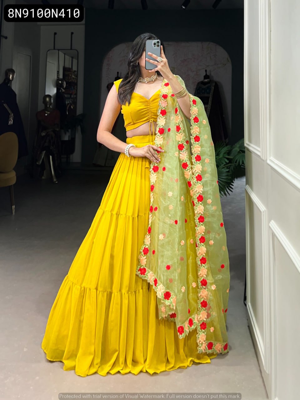 Front full body view of a woman wearing a vibrant TRENDBUY yellow georgette lehenga choli set with a tiered skirt, paired with a light green organza dupatta featuring rich red and peach floral embroidery, model's face obscured by phone.