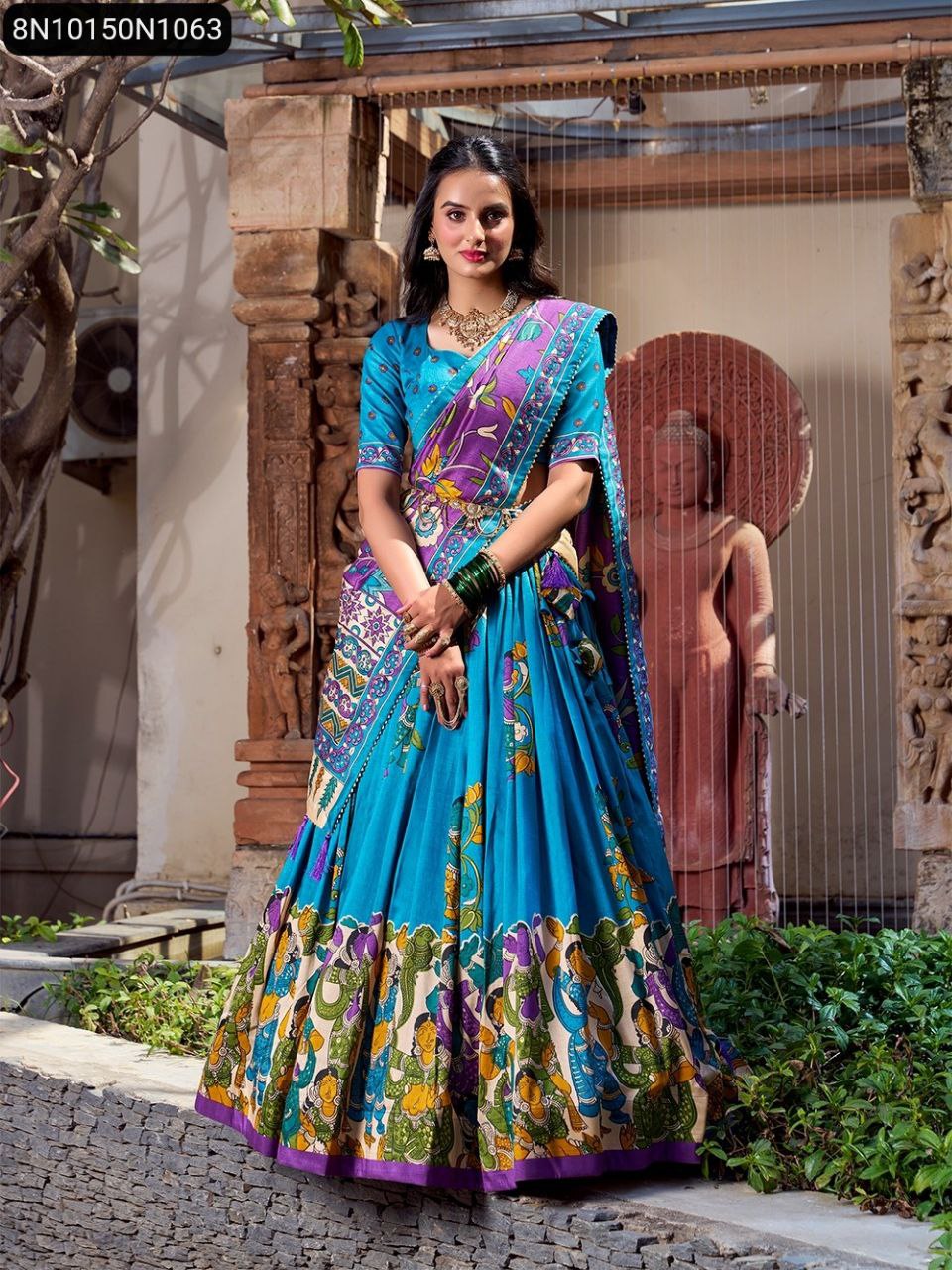 Model in teal blue lehenga choli with purple and green kalamkari print, full front pose, looking directly at the camera, showcasing the complete outfit.
