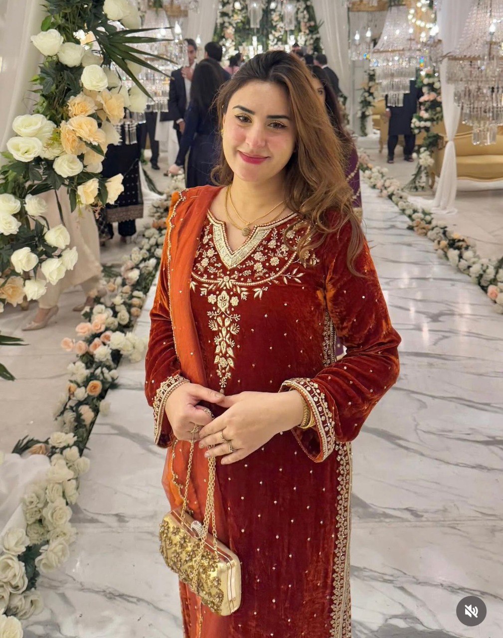 Woman smiling in a rust brown velvet suit, intricately embroidered, standing in a decorated hall.