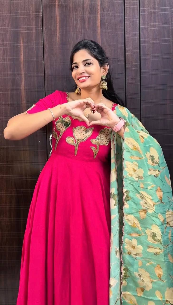 Front view of a woman in a vibrant Hot Pink Vichitra Silk gown with hand-painted Maggam work and a mint green floral dupatta, making a heart gesture.