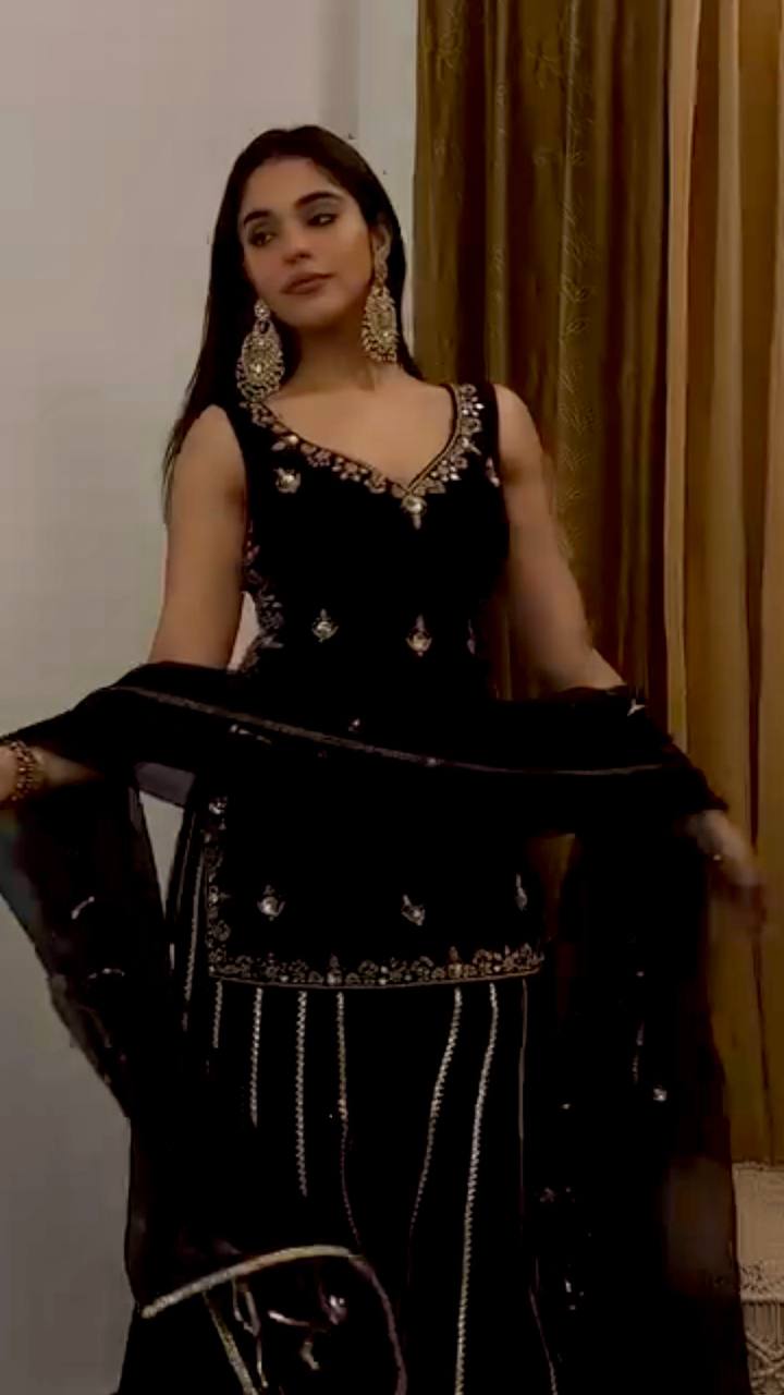 Woman posing elegantly in a black embroidered velvet sharara suit with a matching dupatta.