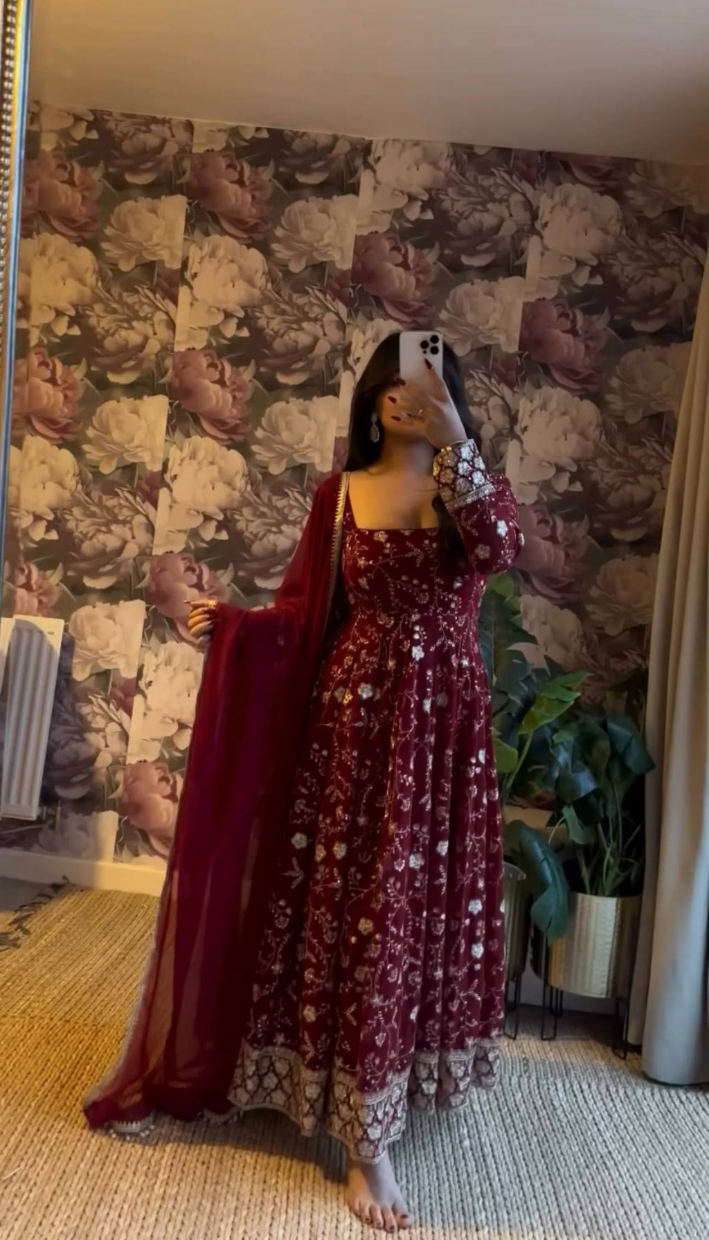 Woman posing in a stunning maroon embroidered Anarkali suit with a phone, showcasing the full design.