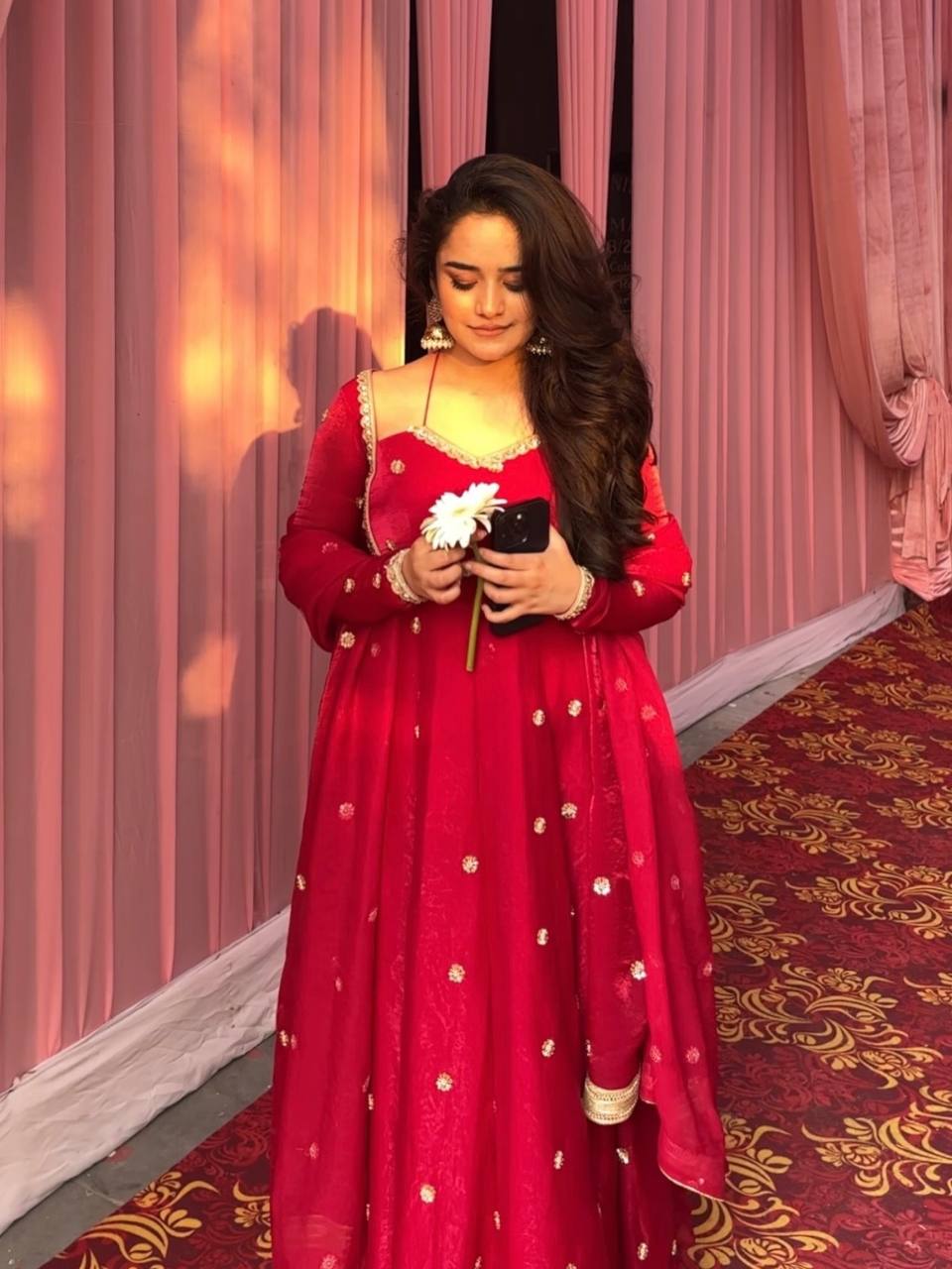 Woman posing gracefully in a red Fendy silk Anarkali suit with gold sequence work, holding a white flower.
