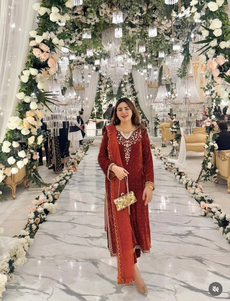Woman standing elegantly in a grand hall adorned with chandeliers and flowers, wearing a Rust Brown velvet embroidered kurta set.