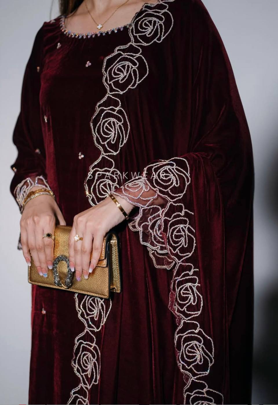 Woman posing elegantly in a burgundy velvet palazzo suit set with intricate white and gold embroidery, holding a golden clutch.