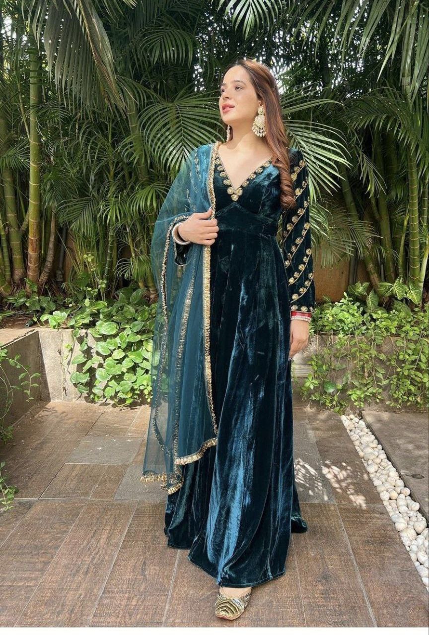 Full body image of a woman posing in a luxurious dark teal velvet party gown with intricate gold embroidery and a matching net dupatta.